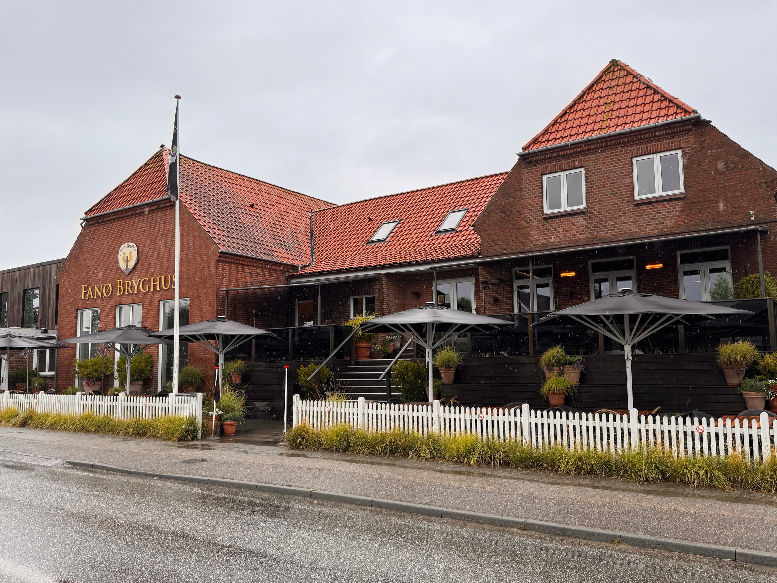 Das Backsteingebäude des Fanø Bryghus mit rotem Ziegeldach und dem Schriftzug samt Wappen an der Giebelwand; davor die Terrasse mit zusammengeklappten Sonnenschirmen und Kübelpflanzen hinter einem weißen Lattenzaun, an einem regennassen Tag.
